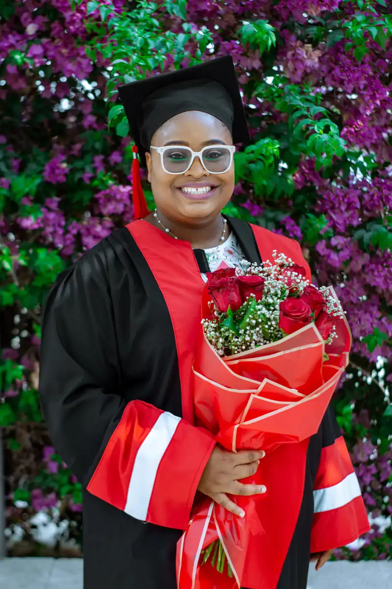 Smiling graduate with glasses holding a bouquet in front of flowering plants